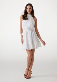 Short white dress with a high neckline, smocked waist, and ruffled hem. Features delicate floral embroidery and a tied fabric belt.
