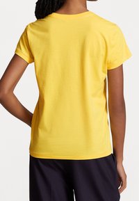 Osoba s pletenými vlasy, oblečená v obyčejném žlutém tričku s krátkým rukávem a tmavých kalhotách, zobrazená zezadu s jednou rukou na boku.