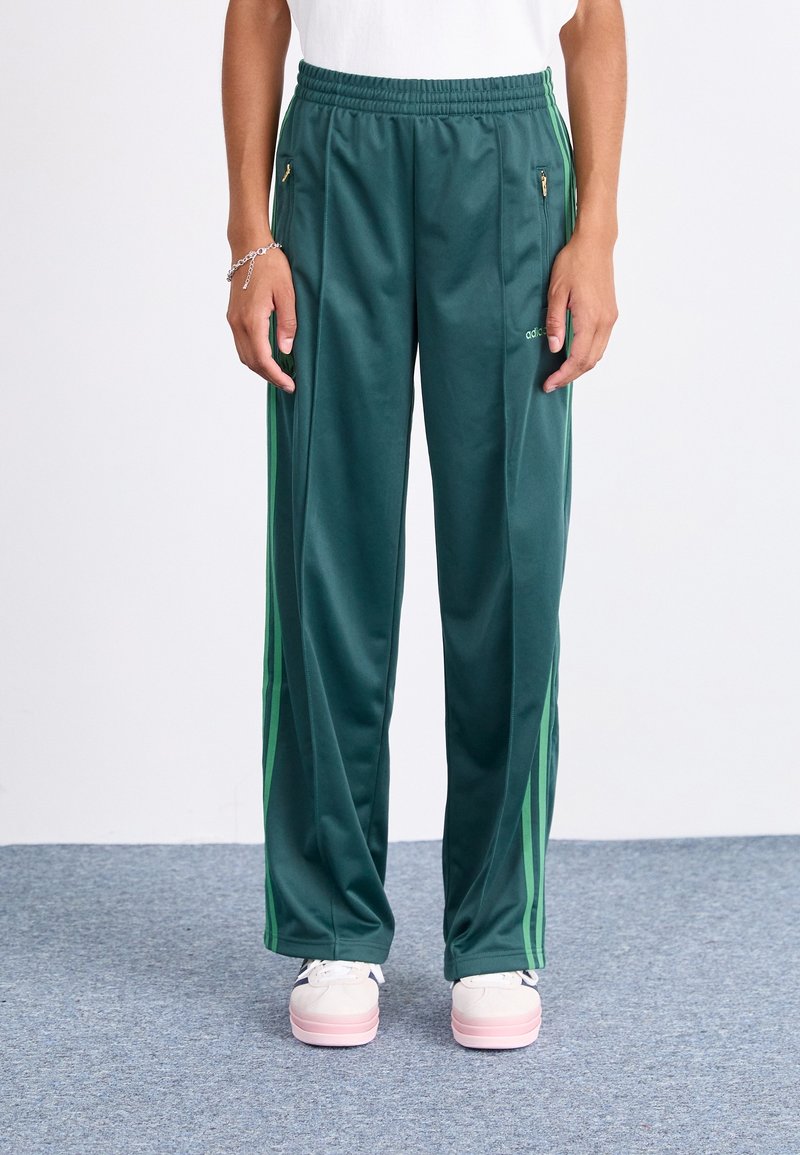 Pantalons de sport verts avec une texture lisse, présentant des bandes latérales vertes, une taille élastique, des poches zippées et un détail de logo brandé.