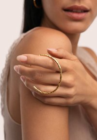 Pulsera de brazalete de oro con textura y diseño abierto, llevada en una mano. Tonos de piel suaves y fondo de tela suave.