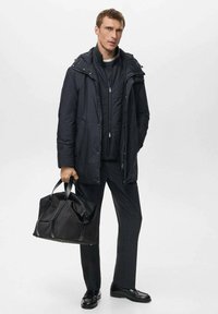 Giacca nera da esterno con cappuccio, indossata sopra un maglione scuro. Tiene una borsa a tracolla nera. Pantaloni scuri e scarpe lucide completano il look.