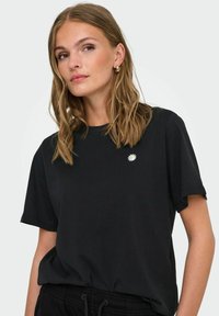 Schwarzes Baumwoll-T-Shirt mit kurzen Ärmeln, das auf der Brust eine kleine weiße Gänseblümchen-Stickerei aufweist. Weiche Textur; lockere Passform.