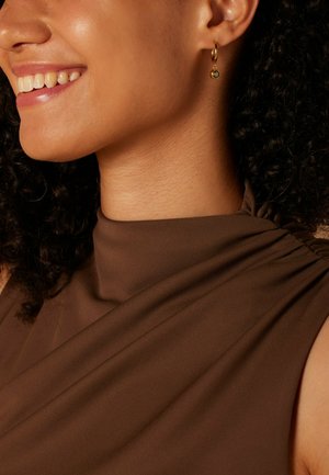 Haut sans manches marron avec un détail froncé à l'épaule, associé à une boucle d'oreille en or avec un accent en pierre précieuse verte.