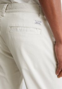Beige Baumwoll-Denim-Hosen mit glatter Textur, die eine Gesäßtasche und ein Etikett mit Text aufweisen. Nahaufnahme von der Seiten- und Rückansicht.
