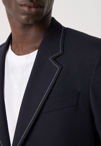 Blazer azul-marinho com costura branca contrastante, bolso no peito, lapelas com cortes, usado sobre uma t-shirt branca. Textura de tecido suave.