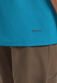 Camicia blu a maniche corte con una texture liscia e un logo che recita "TEXADRI" in basso a sinistra. Abbinata a pantaloni corti leggeri marroni.