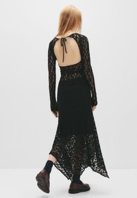 Abito in pizzo nero con maniche lunghe, schiena scoperta fissata da un laccio, orlo irregolare, texture a strati, abbinato a scarpe scure e calze.