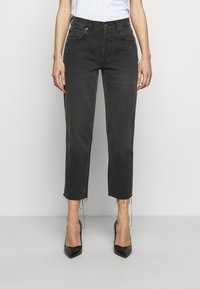 Jeans de mezclilla negra con corte recto, dobladillo sin rematar, diseño de cinco bolsillos y cierre de botones. Combinados con tacones altos negros.