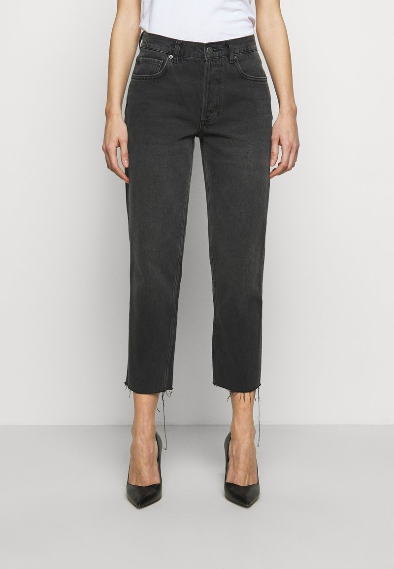 Jeans de mezclilla negra con corte recto, dobladillo sin rematar, diseño de cinco bolsillos y cierre de botones. Combinados con tacones altos negros.