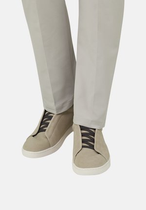 Sneakers in camoscio beige chiaro con lacci neri indossate con pantaloni dritti di colore chiaro su sfondo bianco.