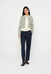 Πλεκτή κρεμ ζακέτα με μαύρες ρίγες, χρυσά κουμπιά, μπεζ τοπ, ναυτικό μπλε παντελόνι και λαμπερά μαύρα παπούτσια με τακούνι, συνδυασμένα με λευκή τσάντα.