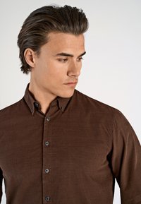 Bruine geknoopte overhemd met een textuurstof, kraag en donkere knopen, slim fit ontwerp, getoond op een model dat iets zijwaarts kijkt.