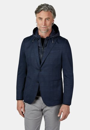 Mittelalter Mann mit grauen Haaren, der einen marineblauen Blazer über einer schwarzen Kapuzen-Zip-Jacke und hellgrauen Hosen trägt, vor weißem Hintergrund stehend.