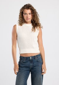 Cream knitted sleeveless top with ribbed texture and open stitch pattern, paired with blue high-waisted jeans.