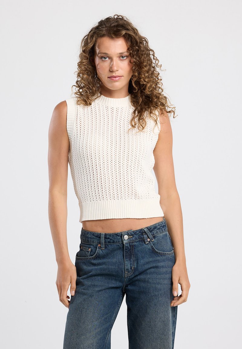 Cream knitted sleeveless top with ribbed texture and open stitch pattern, paired with blue high-waisted jeans.