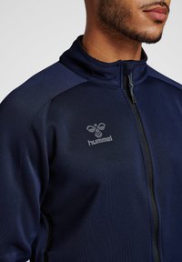 Veste zippée bleu marine en tissu lisse, dotée d'un col montant et d'un logo gris subtil sur la poitrine gauche. La texture semble douce.