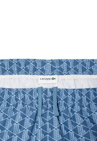 Bañadores de color azul con un diseño geométrico. Cuentan con una cinturilla elástica con cordones y una etiqueta con el logo de Lacoste.