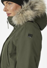 Parka verde oliva con capucha forrada de piel sintética, tejido texturizado, cierre frontal de cremallera y parche de marca en la manga. Tiene un diseño ajustado.