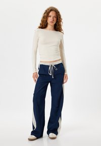 Pantalons larges bleu marine avec des rayures blanches sur les côtés et une taille élastique dotée d'un cordon de serrage. Associés à un haut beige à manches longues.