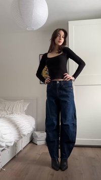 Νεαρή γυναίκα ποζάρει με τα χέρια στη μέση φοράει μαύρο μακρυμάνικο μπλουζάκι, σκούρα μπλε παντελόνι φαρδιάς γραμμής και μαύρα μποτάκια στον αστράγαλο στο υπνοδωμάτιο.