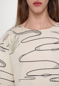 Løse plagg med en kremfarget bakgrunn, som har svarte, svingende linjer og et grønt botanisk design. Myk stofftekstur er synlig.