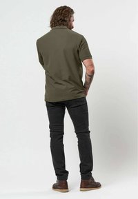 Homme avec des tatouages portant un polo vert olive, un jean noir et des chaussures en cuir marron, debout de dos devant un fond uni.
