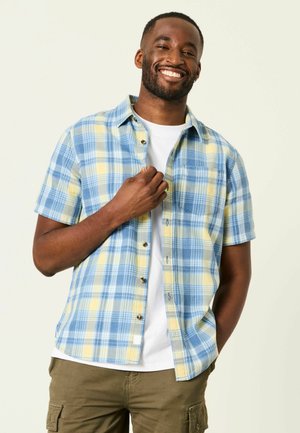 Lächelnder Mann in einem blau-gelb karierten kurzärmeligen Hemd über einem weißen T-Shirt, steht mit einer Hand in der Tasche und richtet mit der anderen das Hemd.