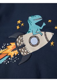 Blauwe dinosaurus die op een beige raket met zwarte vinnen rijdt, vlammen komen uit de achterkant, omringd door kleine gele sterren op een donkerblauwe achtergrond.