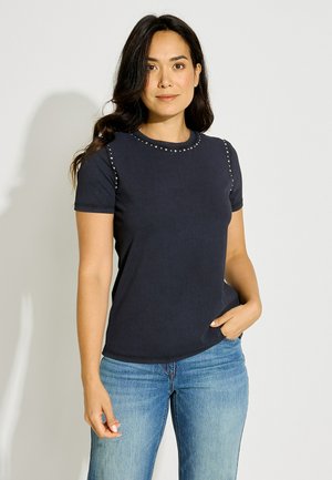 Mujer con cabello largo y oscuro, que lleva una camiseta negra de manga corta con tachuelas y vaqueros azules, de pie con una mano en el bolsillo frente a un fondo liso.