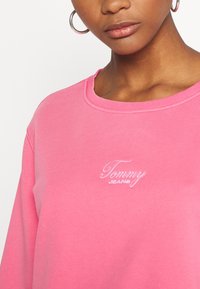 Mujer con arete de aro plateado y sudadera rosa con el logo bordado en blanco "Tommy Jeans" en el pecho, vista en ángulo.