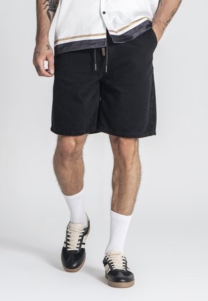 Schwarze Jeansshorts mit entspannter Passform, ausgestattet mit einem normalen Taillenbund und einer Kordel, kombiniert mit weißen Socken und schwarzen Turnschuhen.