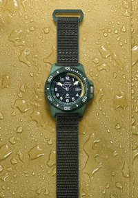 Ρολόι Timex Expedition με ηλιακή ενέργεια και μαύρο υφασμάτινο λουράκι σε υγρή, υφή κίτρινη επιφάνεια που δείχνει την ώρα 10:08 και ημερομηνία.