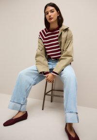 Giacca oversize color kaki, maglione a righe bordeaux e bianche, jeans chiari a vita alta e ballerine bordeaux, seduta su uno sgabello di legno.