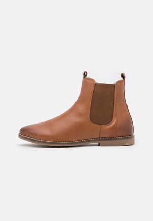 Botte Chelsea en cuir marron avec panneaux latéraux élastiques, bout arrondi et talon bas. Hauteur cheville avec languettes pour un enfilage facile.