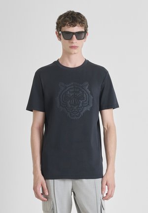 Joven con gafas de sol negras, camiseta negra con un rostro de tigre en relieve y pantalones cargo gris claro contra un fondo claro y liso.