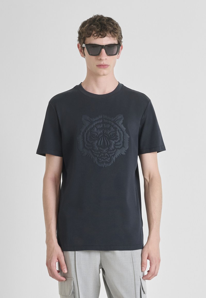 Mladić nosi crne naočale za sunce, crnu majicu s reljefnim motivom tigra i svijetlosive cargo hlače na jednoličnoj svijetloj pozadini.