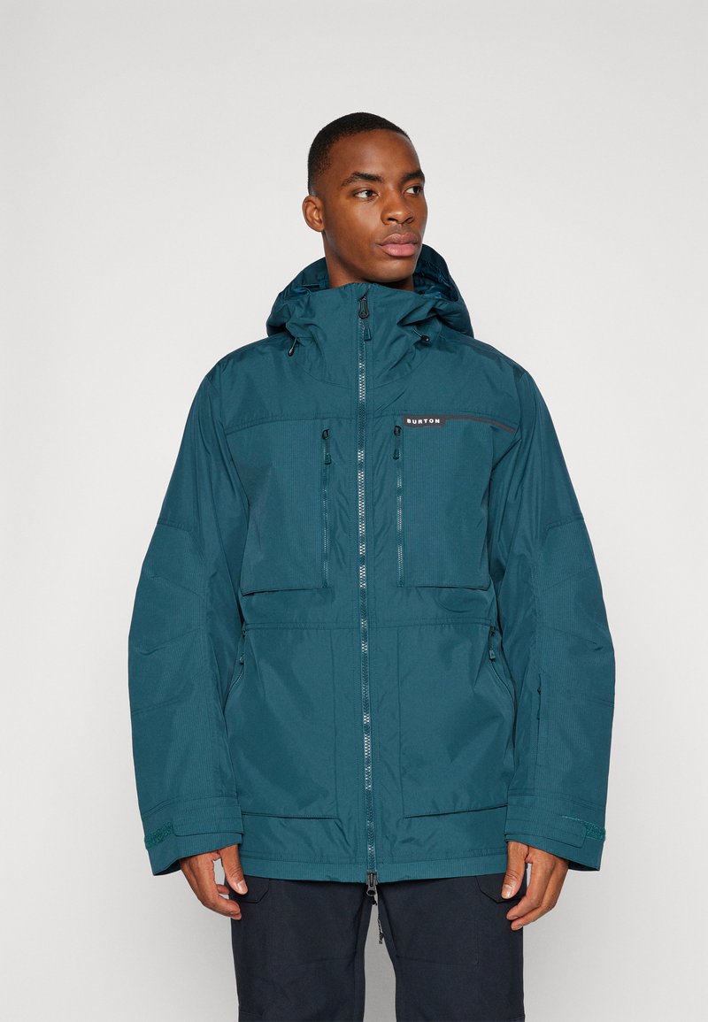 Veste imperméable bleu sarcelle avec capuche, dotée de deux poches avant zippées, de poignets ajustables et d'un détail de logo sur la poitrine.