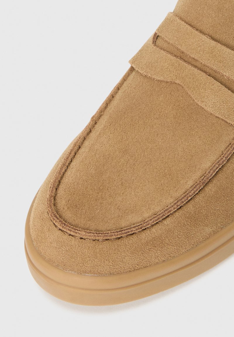 Zapato de ante color beige, tipo mocasín, con punta redondeada, que presenta un empeine cosido y una suela de goma flexible. Textura suave y diseño minimalista.