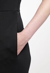 Vestido negro con un diseño ajustado, que presenta un bolsillo lateral. La tela parece suave y ligeramente elástica. Una mano está apoyada en el bolsillo.