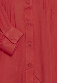 Blusa roja de botones con una tela texturizada, que presenta una manga plisada y seis botones cubiertos de tela a lo largo del frente para el cierre.