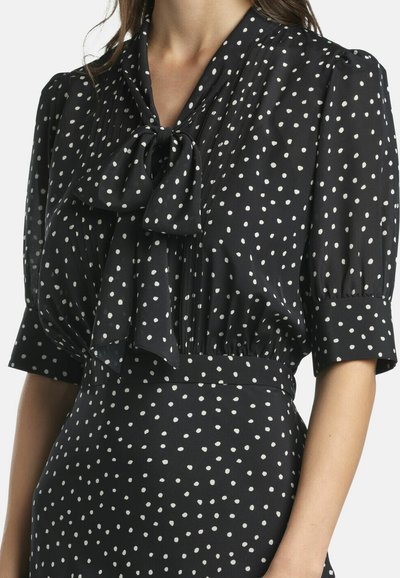 Črna bluza z belimi pikami, z mašno na ovratniku, kratkimi napihnjenimi rokavi in zbirano teksturo okoli pasu.