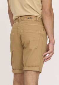Shorts khaki avec un ourlet roulé, fabriqués en tissu coton robuste. Comprend des poches arrière et une étiquette de marque visible sur la ceinture.