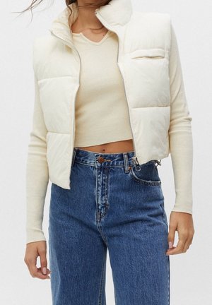 Femme portant une doudoune courte crème par-dessus un crop top côtelé beige à manches longues, avec un jean en denim bleu taille haute, debout devant un fond uni.