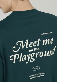 T-shirt vert foncé en coton doux, avec un texte blanc : "Rencontrons-nous sur le terrain de jeu", avec une coupe décontractée et un col rond.