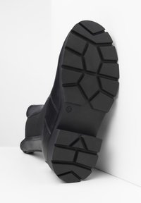 Botín negro con una suela de goma gruesa y texturizada que presenta un patrón de tracción hexagonal. Parte superior compuesta de materiales de cuero liso y mate.