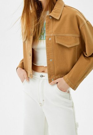 Kvinde iført en kort beige jakke over en hvid crop top og hvide jeans med høj talje, hænderne i lommerne, på en ensfarvet baggrund.
