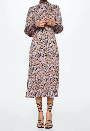 Femme portant une robe fleurie à manches longues et col haut, longueur midi, associée à des sandales noires à talons et à lanières.