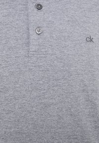 Siva polo majica od mekog materijala, s dvorednim dugmadi i malim izvezenim "ck" logom s lijeve strane.
