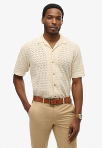 Kortærmet, cremefarvet skjorte med knapper og et tekstureret zigzag-mønster, parret med beige bukser og et brunt læderbælte.