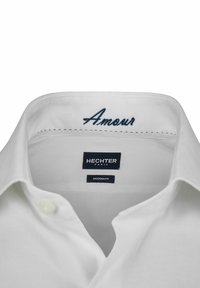 Weißes Hemdkragen mit blauer bestickter Aufschrift "Amour", versehen mit einem Markenetikett: "HECHTER PARIS" und "MODERN FIT". Glatte Stofftextur.
